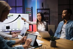 Four multi ethnic economists sitting at office with various gadgets and analysing financial growth in worldwide economy. Male and female partners having business meeting during evening time.